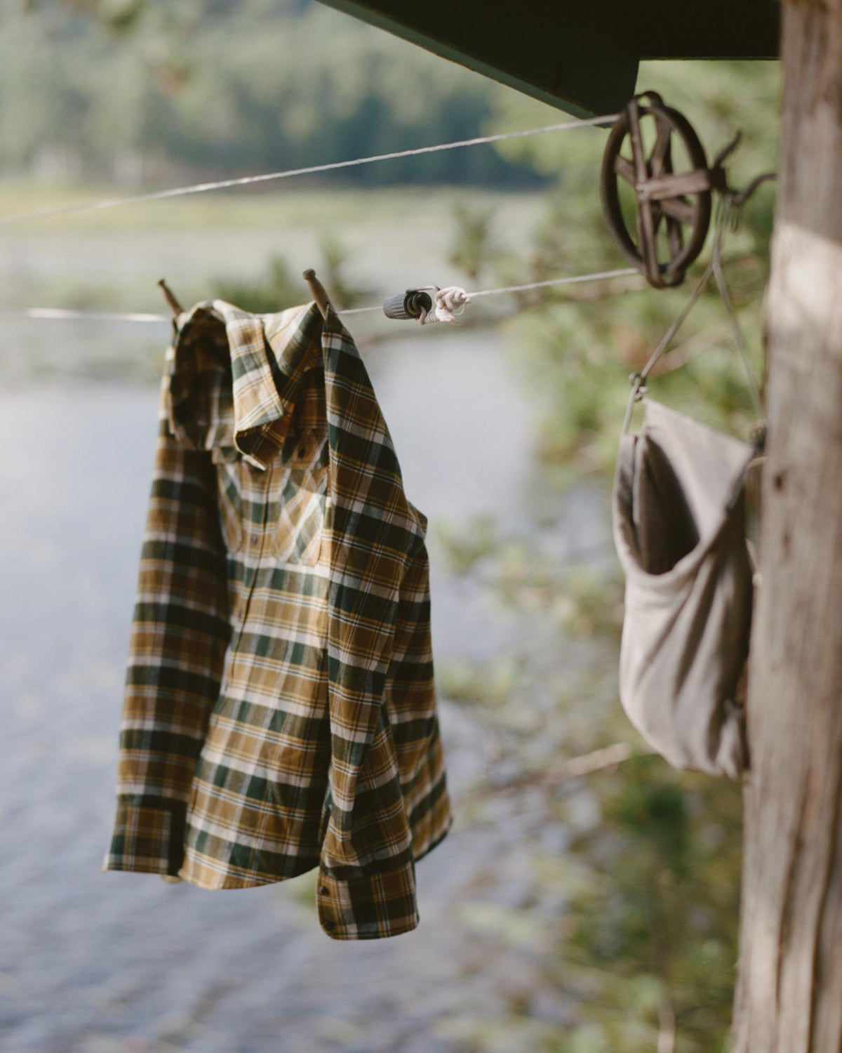 A plaid shirt and a canvas bag hang from a clothesline outdoors near water, with trees in the background.