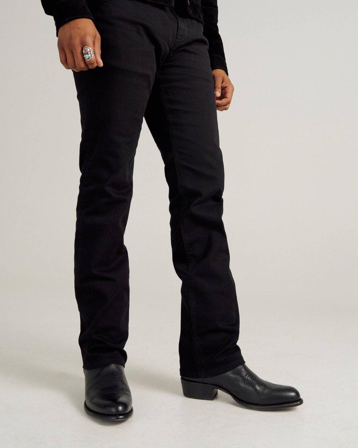 Person wearing black pants, black boots, and a ring, standing against a plain light background. Only the lower body is visible.