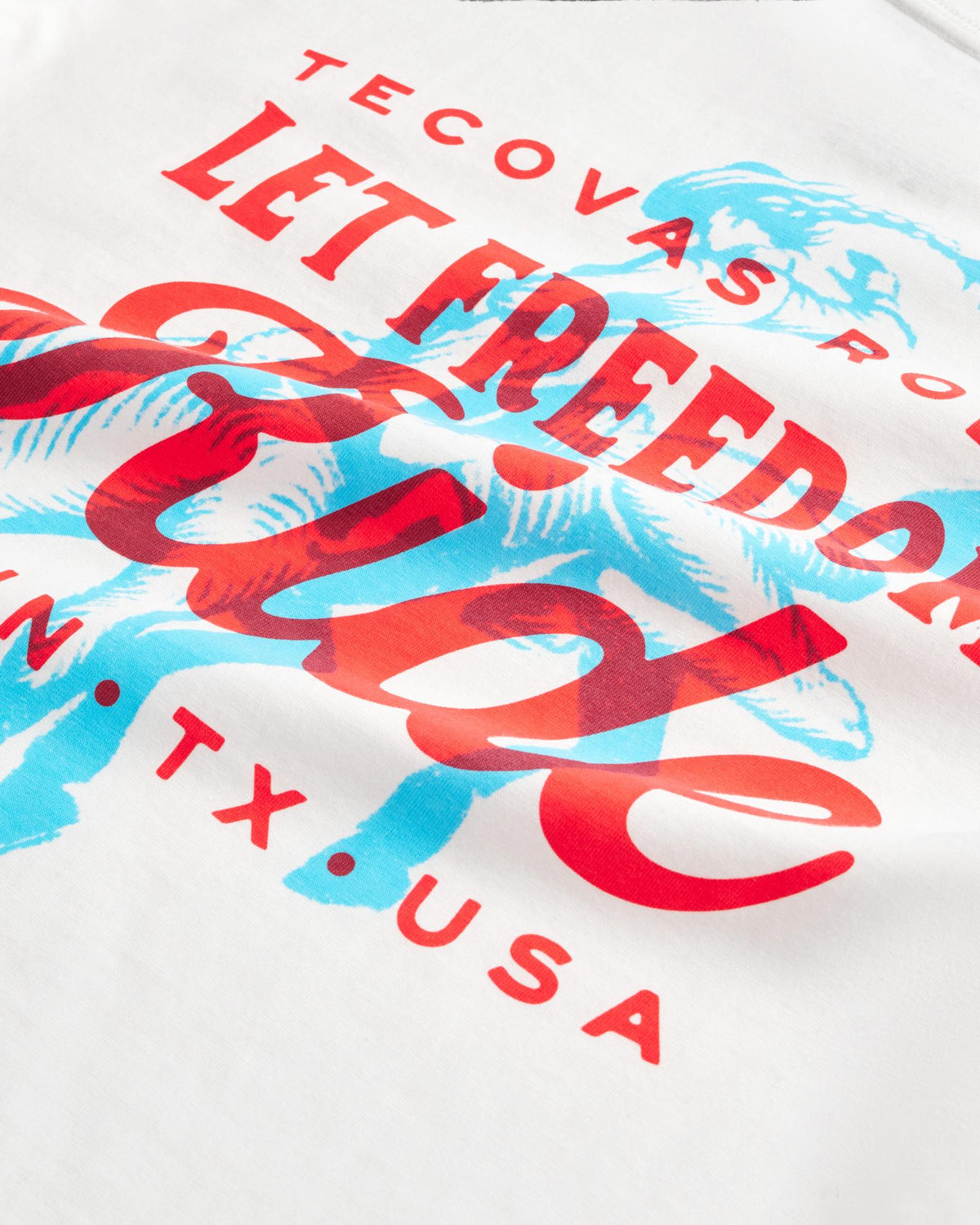 White t-shirt with red and blue graphic text reading “Tecovas presents Let Freedom Ride” and “ATX, TX, USA,” featuring a cowboy on a horse illustration.