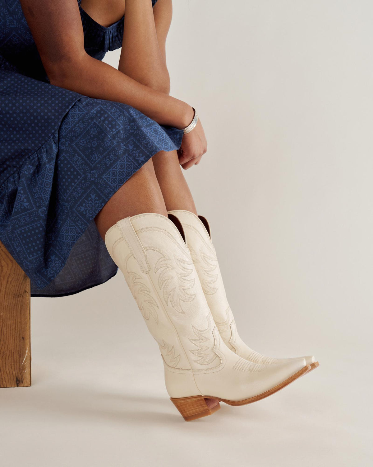 Person in a blue patterned dress sits on a wooden bench, wearing cream-colored cowboy boots with decorative stitching and wooden heels.