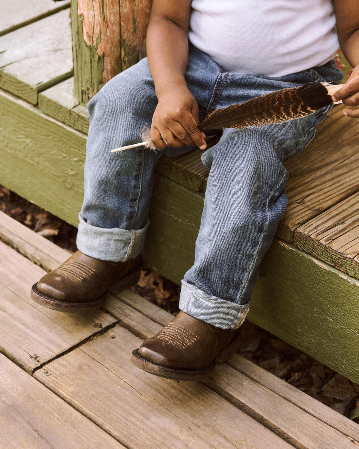A child wearing jeans and boots sits on green wooden steps, holding a large feather.