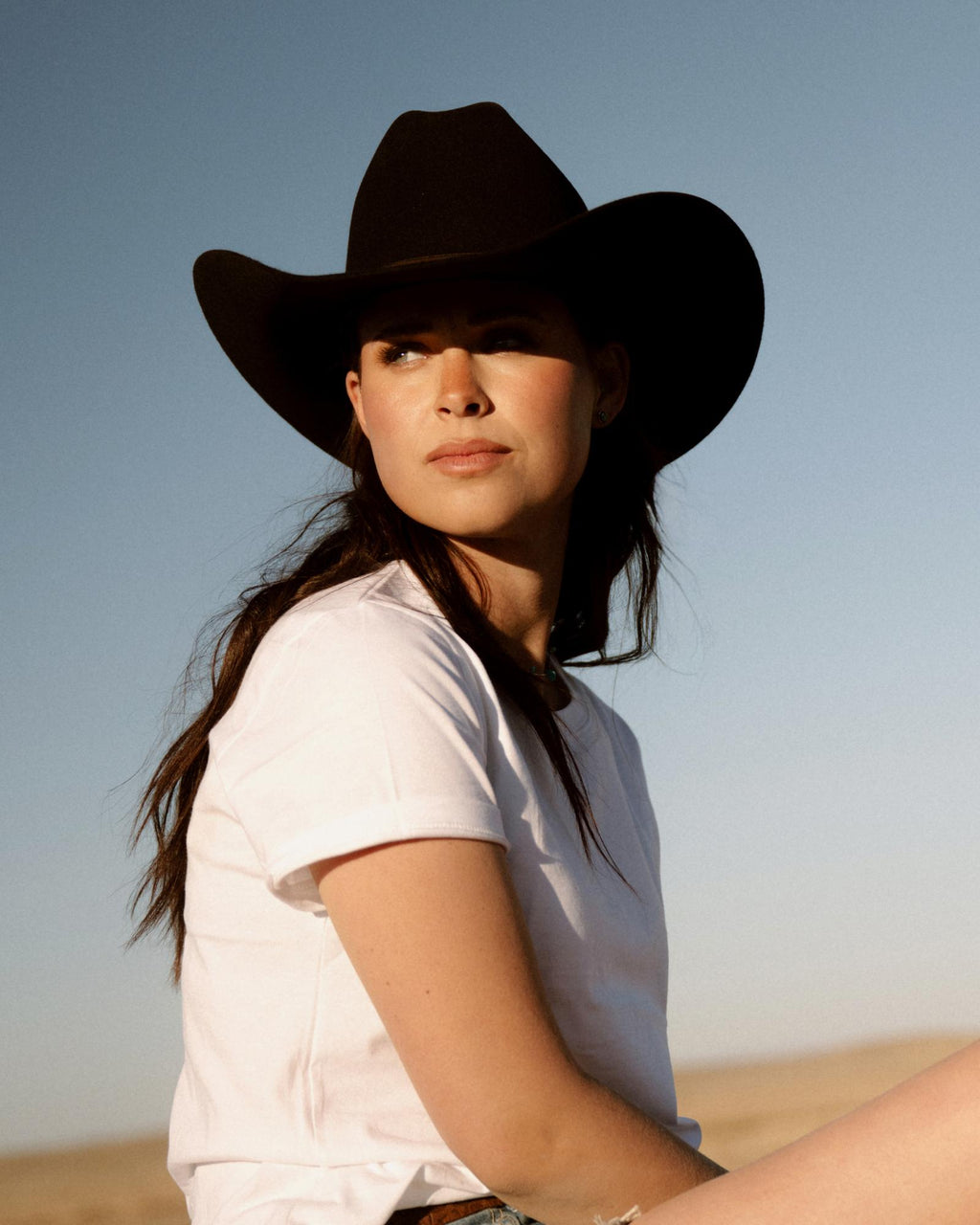 Woman in a white t-shirt and black cowboy hat sits outdoors, looking to the side, with a clear sky and desert landscape in the background.