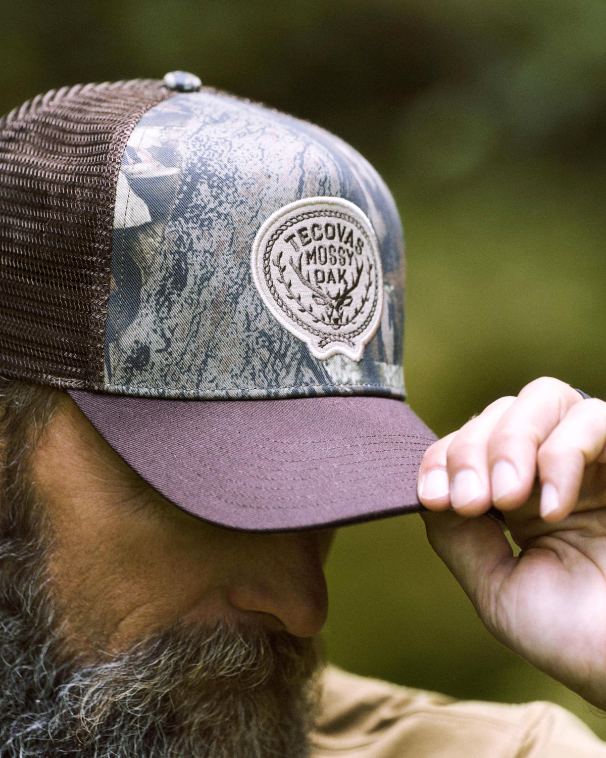 A person with a beard wears a camouflage mesh cap with a “Tecovas Mossy Oak” patch, holding the brim with their hand.