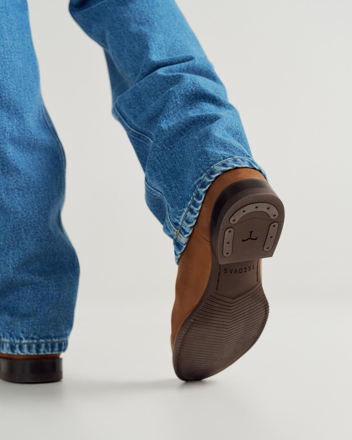 The Glen in Khaki Cowhide. Person wearing blue jeans and brown leather shoes with a visible rubber sole and metal plate, walking forward on a light-colored surface, pairs the look with a crisp khaki shirt for casual sophistication.