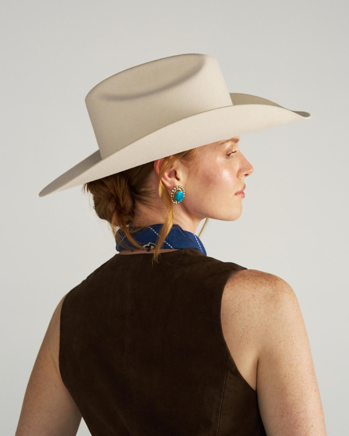 A woman in a beige wide-brimmed hat and dark vest faces sideways. She has a braid, wears a blue scarf, and a turquoise earring.