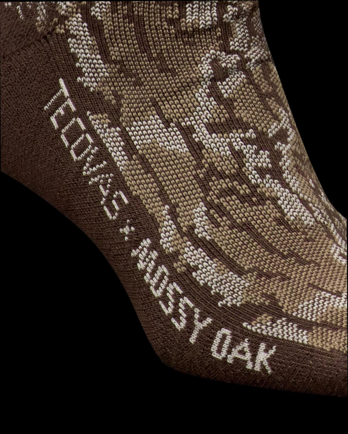 Close-up of a brown sock with a camouflage pattern and the text "TECOVAS x MOSSY OAK" knitted into the fabric near the ankle.