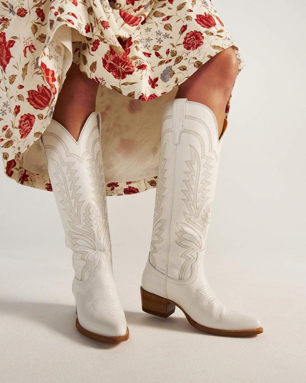 The Abby Wide Calf in Snow Cowhide. Person wearing a floral skirt and white Snow Cowhide embroidered cowboy boots with a wooden heel, standing on a light background.