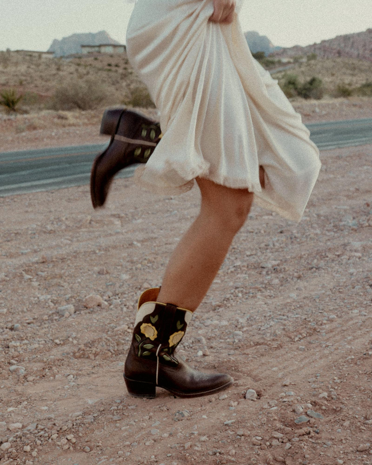 The Jolene in Truffle Cowhide. A person in a white dress and embroidered truffle cowhide cowboy boots walks on a gravel road in a desert landscape.