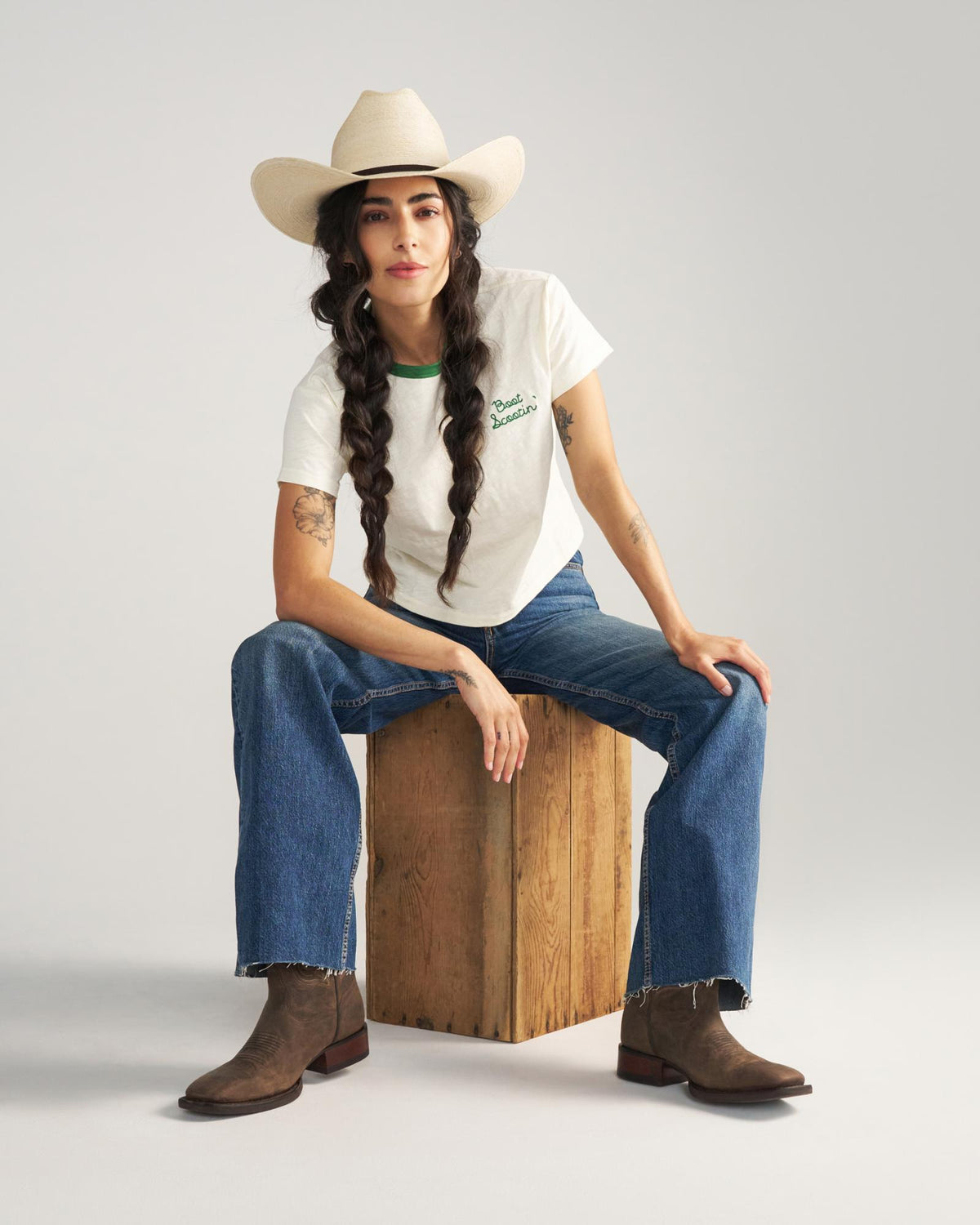 The Tess in Cafe Goat. A person wearing a cowboy hat, white t-shirt, blue jeans, and brown boots sits on a wooden box against a plain background, evoking the relaxed charm of Cafe Goat.