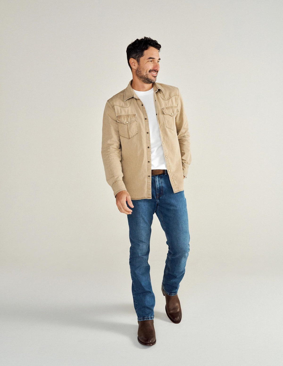 Man in a beige button-up shirt over a white T-shirt, blue jeans, and brown boots, standing with a smile against a plain background.