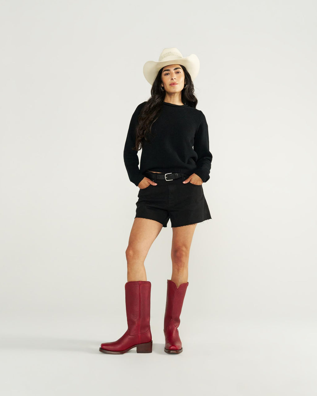 The Charlie in Scarlet Cowhide. Woman in a white cowboy hat, black sweater, black shorts, and striking Scarlet Cowhide red cowboy boots stands against a plain white background.