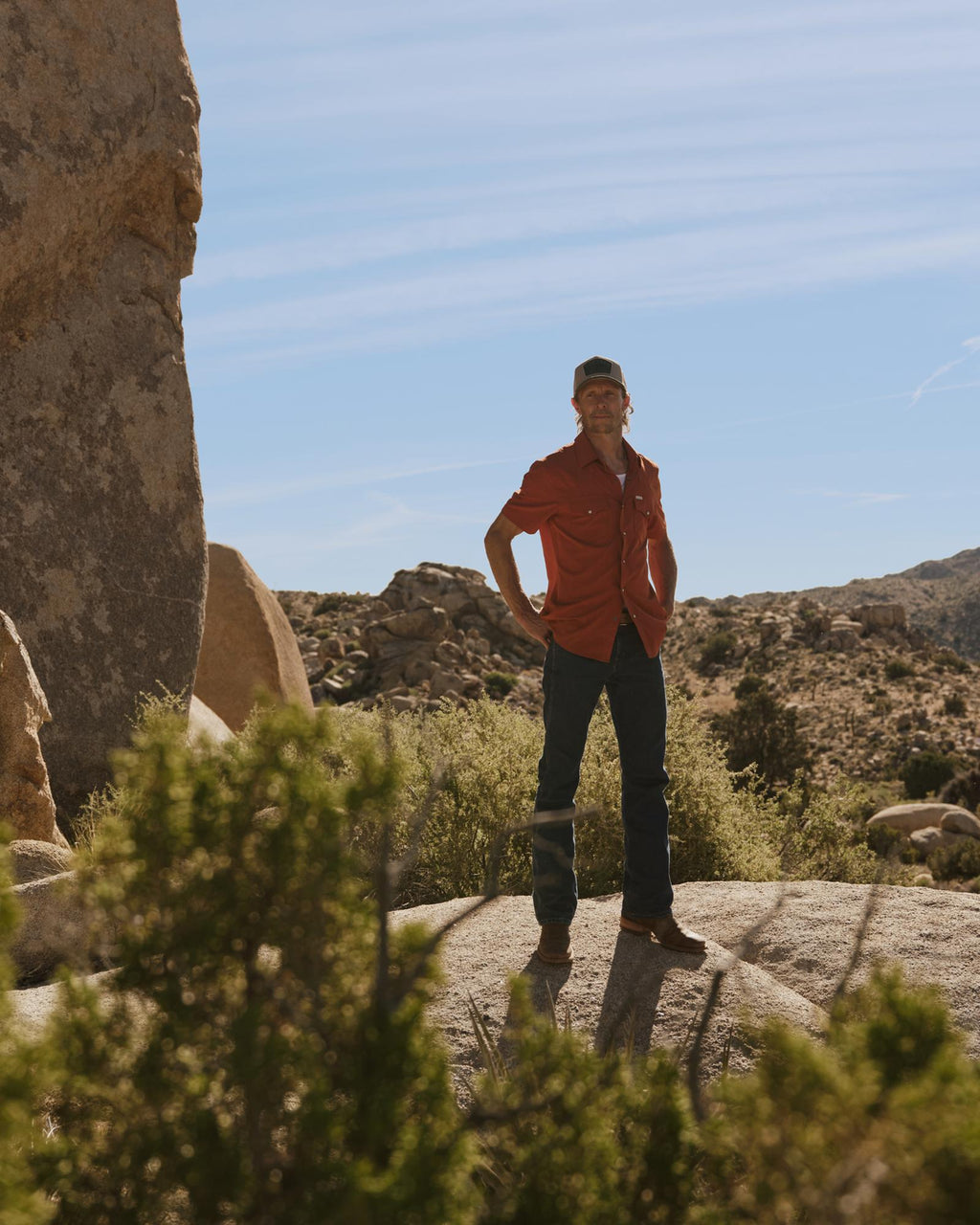 A man in a red shirt and jeans stands on a rocky landscape with desert vegetation and large boulders under a blue sky.
