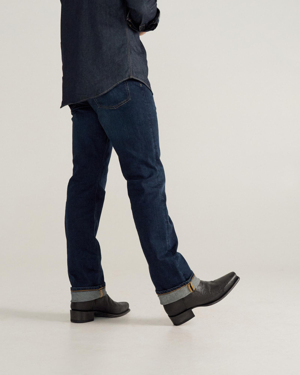 Person wearing dark blue jeans, a dark shirt, and black boots is walking, shown from the shoulders down against a plain background.