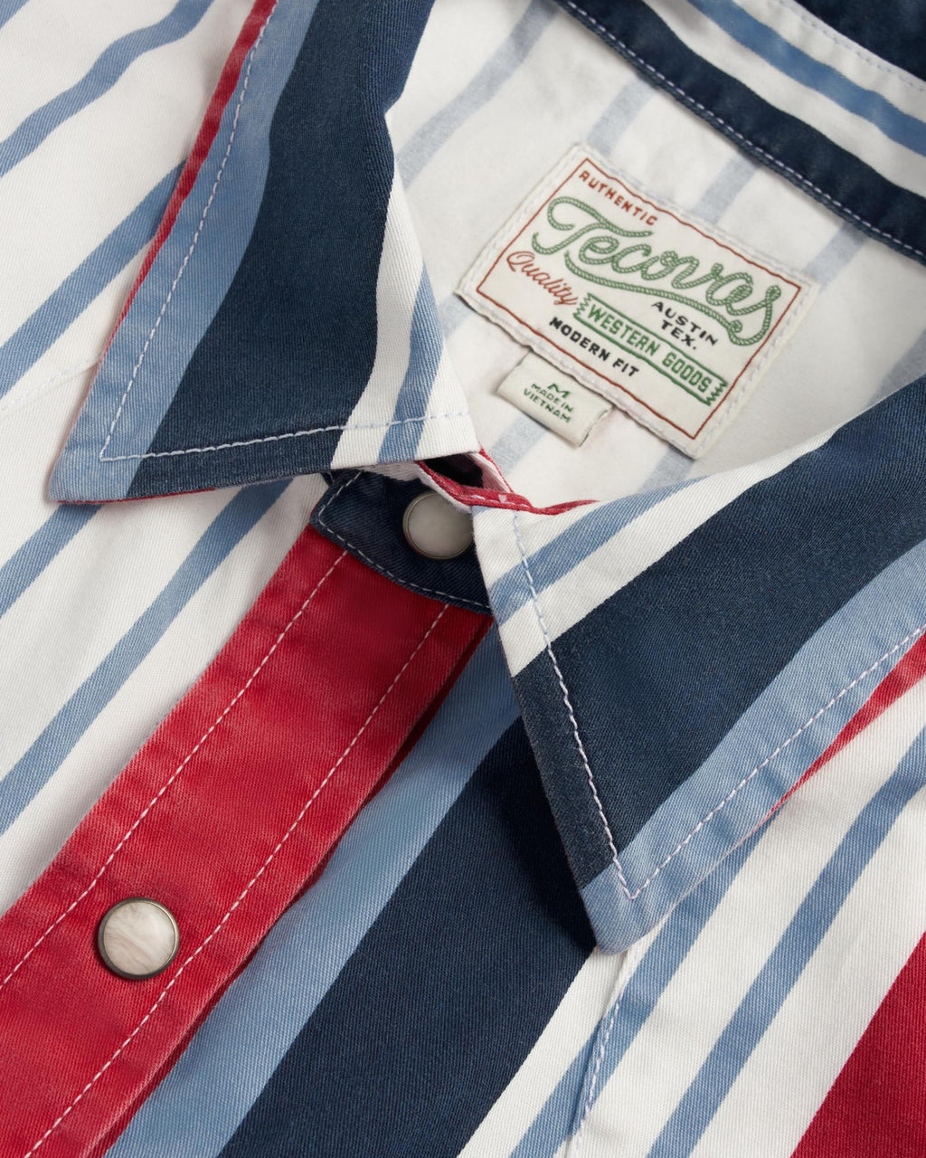 Close-up of a collared shirt with vertical red, white, and blue stripes, snap buttons, and a sewn label that reads "Genuine Western Goods, Modern Fit.
