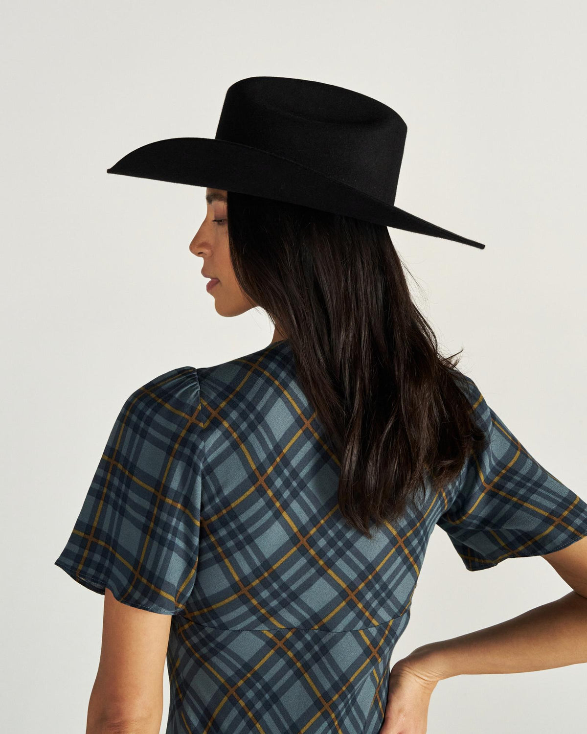 Woman in a plaid dress and black wide-brim hat stands facing away, showing her profile and long dark hair against a plain background.