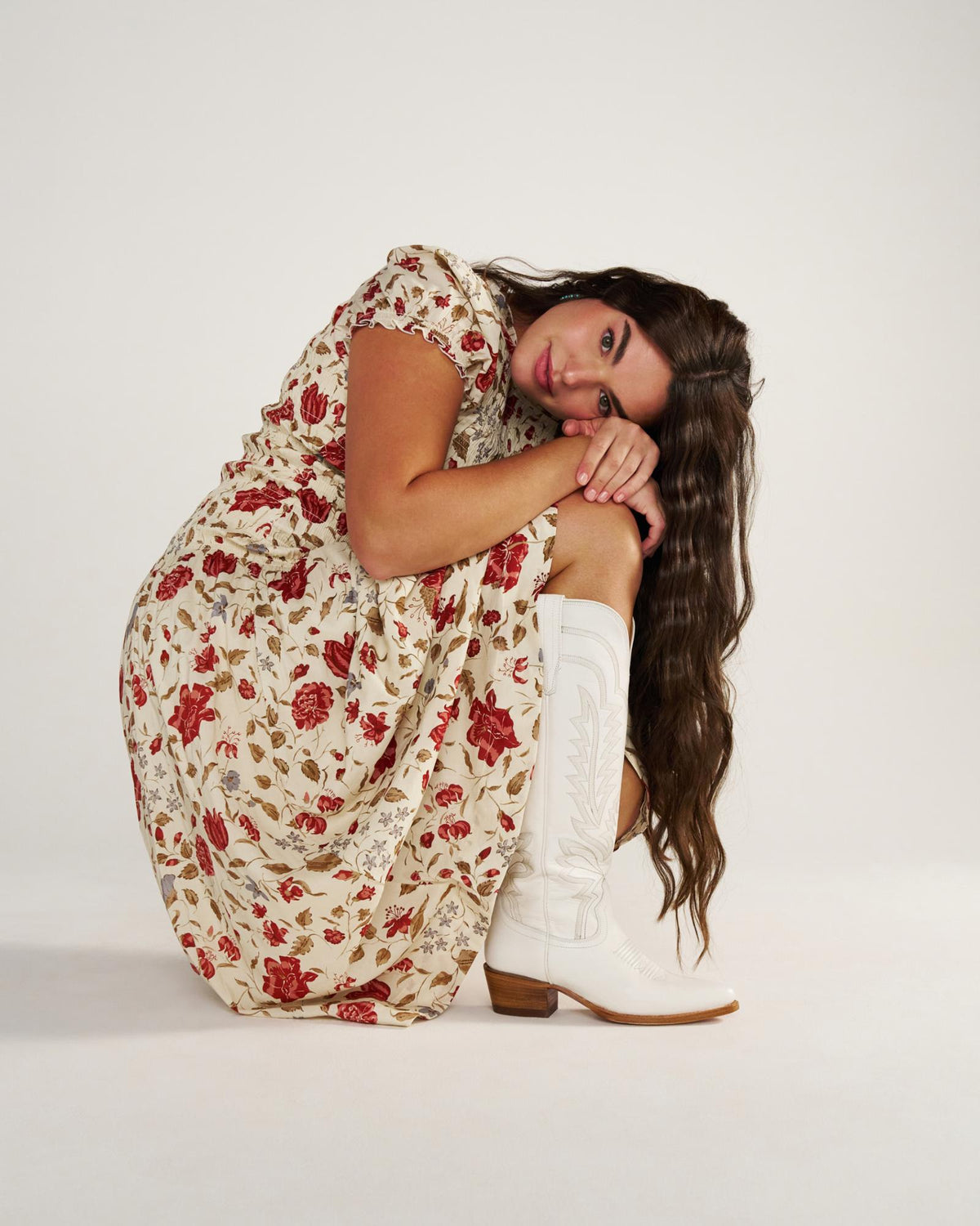 The Abby Wide Calf in Snow Cowhide. A woman in a floral dress and white cowboy boots sits on the ground with her knees to her chest, looking at the camera against a plain white background, her boots featuring a hint of cowhide for a chic Western touch.