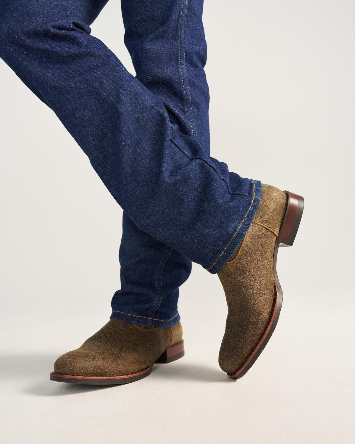 The Glen in Dustwood Roughout. Person wearing blue jeans and brown Dustwood suede boots stands with legs crossed against a plain background.