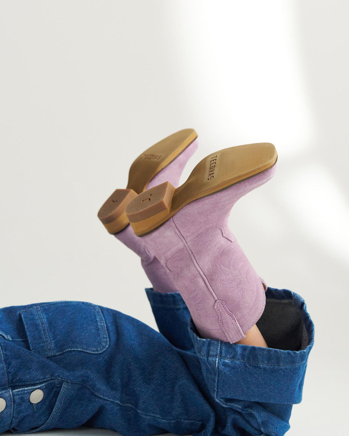 A person wearing denim pants and light purple heeled boots with tan soles is lying down with their legs raised.