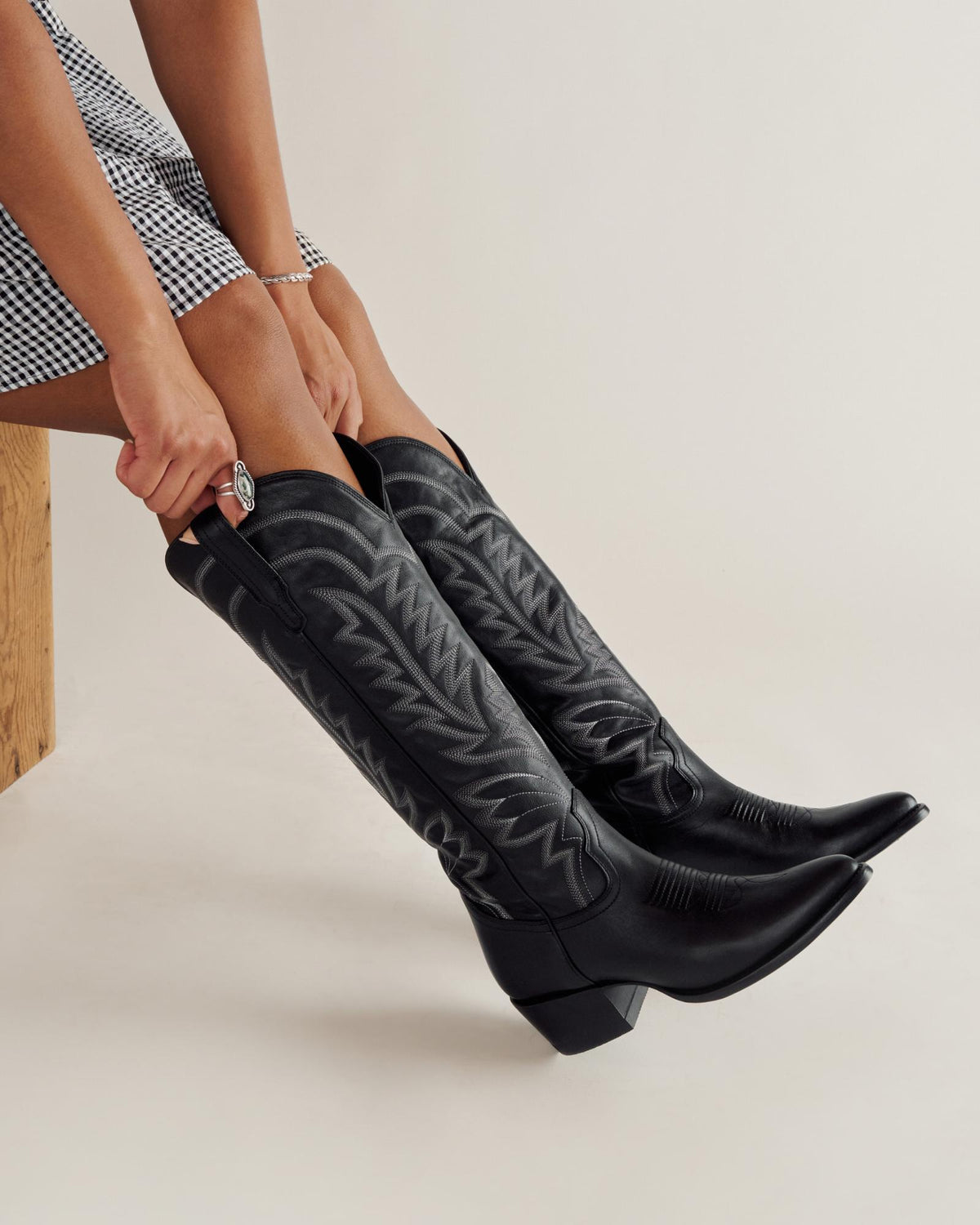 A person wearing a black-and-white checkered skirt is putting on tall black cowboy boots with decorative stitching.