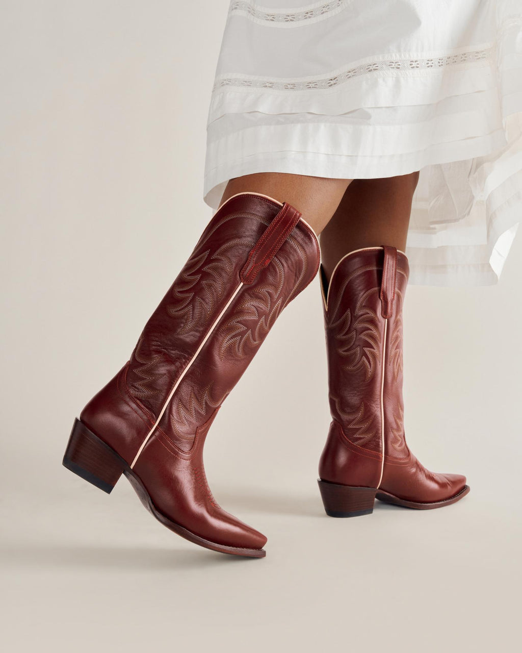 A person wearing a white dress and brown leather cowboy boots with decorative stitching and a low heel, standing against a plain background.