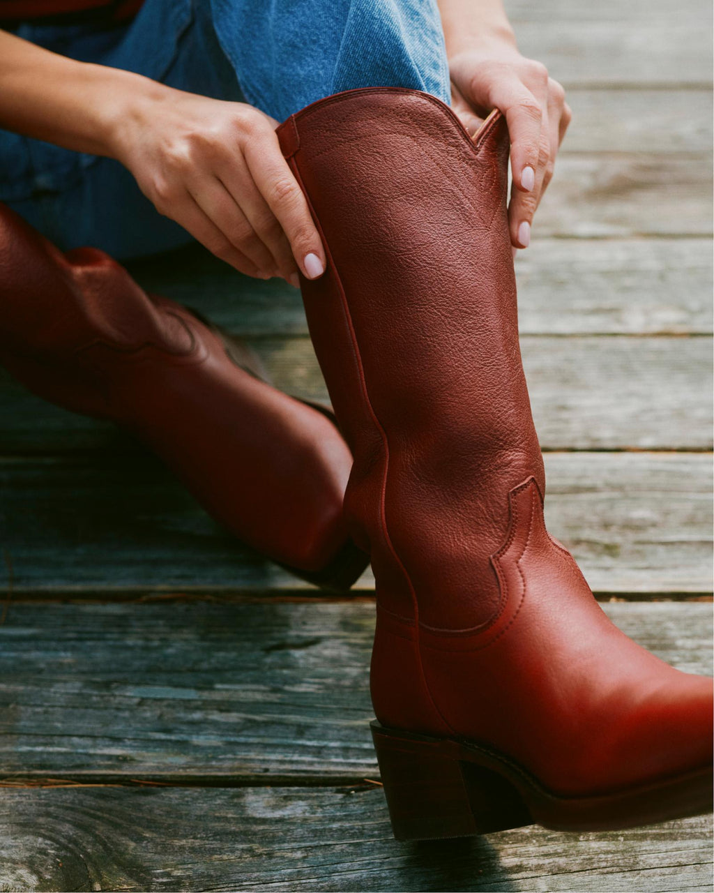 The Charlie in Scarlet Cowhide. Person wearing blue jeans puts on tall brown Scarlet Cowhide cowboy boots while sitting on a wooden surface.