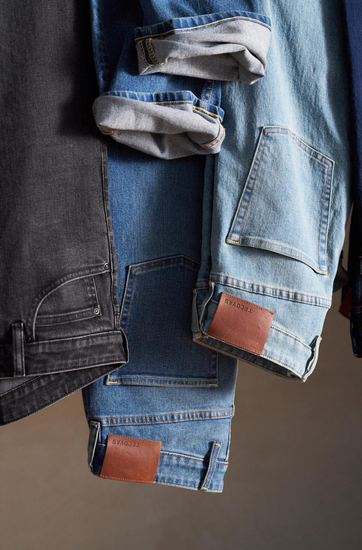 Three pairs of jeans, including black, medium blue, and light blue, are hung side by side with visible brown leather patches and rolled-up cuffs.