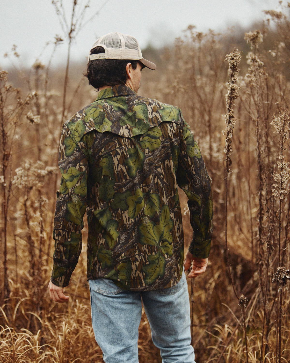 A person wearing a camouflage shirt, blue jeans, and a cap stands in a field of tall, dry grass, facing away from the camera.