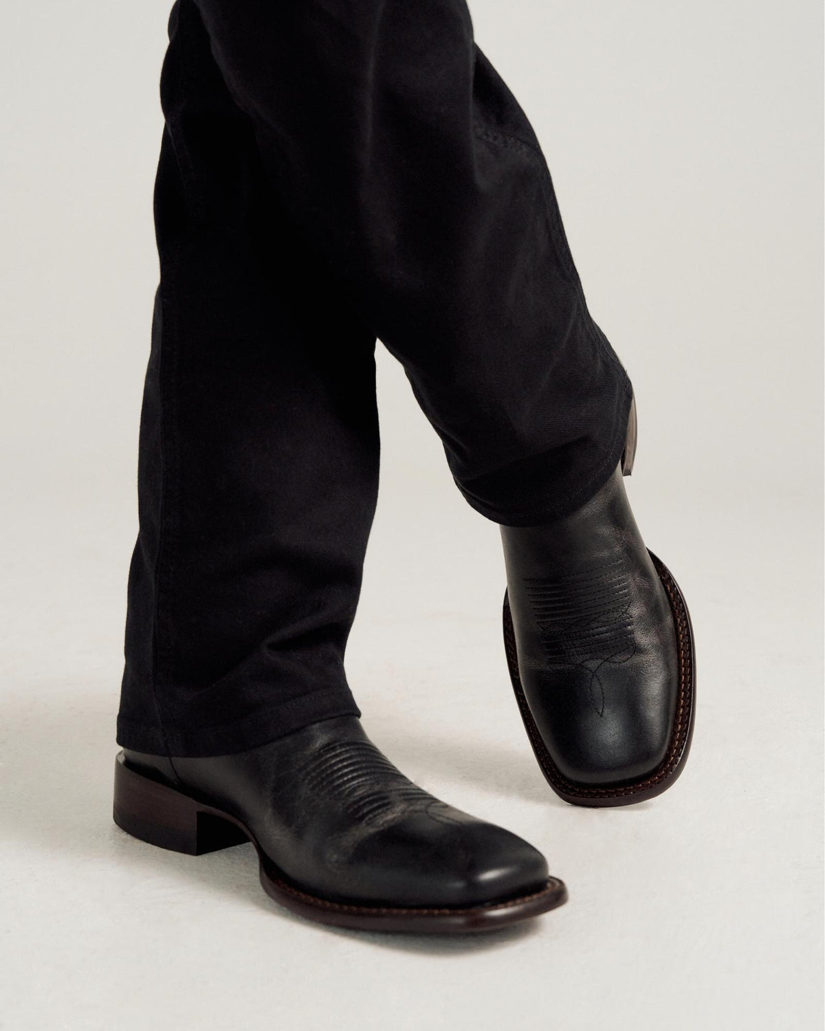 Person wearing black leather cowboy boots and black pants, standing with legs crossed on a light-colored surface.