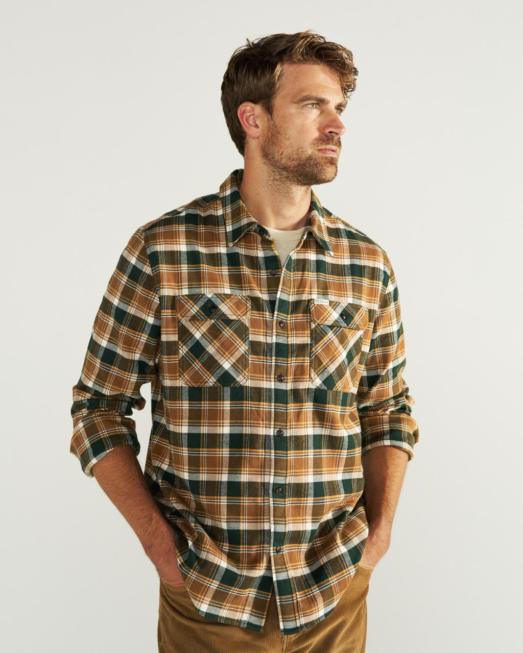 Man wearing a brown, green, and white plaid button-up shirt over a light t-shirt, standing with hands in pockets against a plain light background.