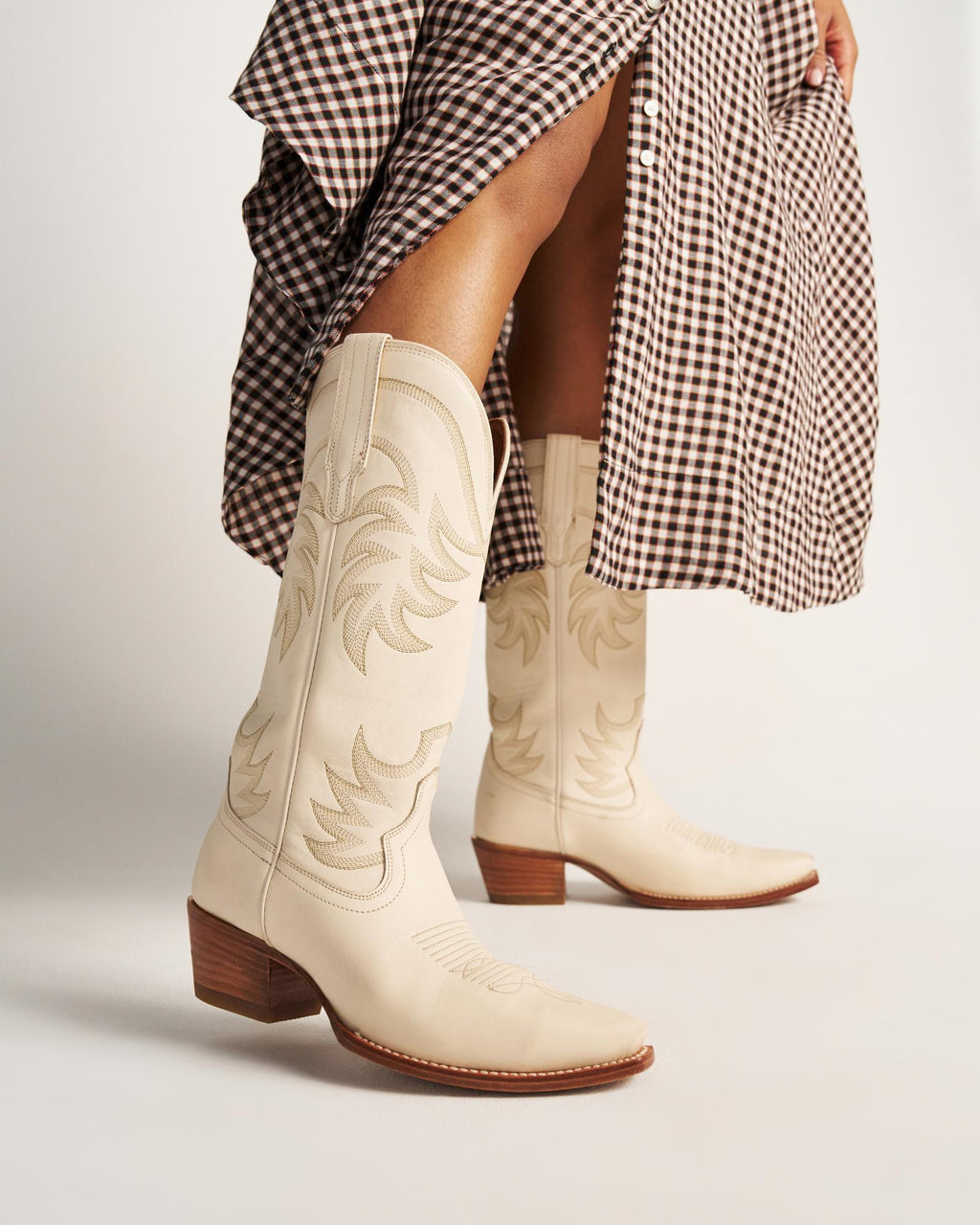 The Annie in Bone Cowhide. A person wearing bone cowhide cowboy boots and a brown checkered skirt stands against a plain background.