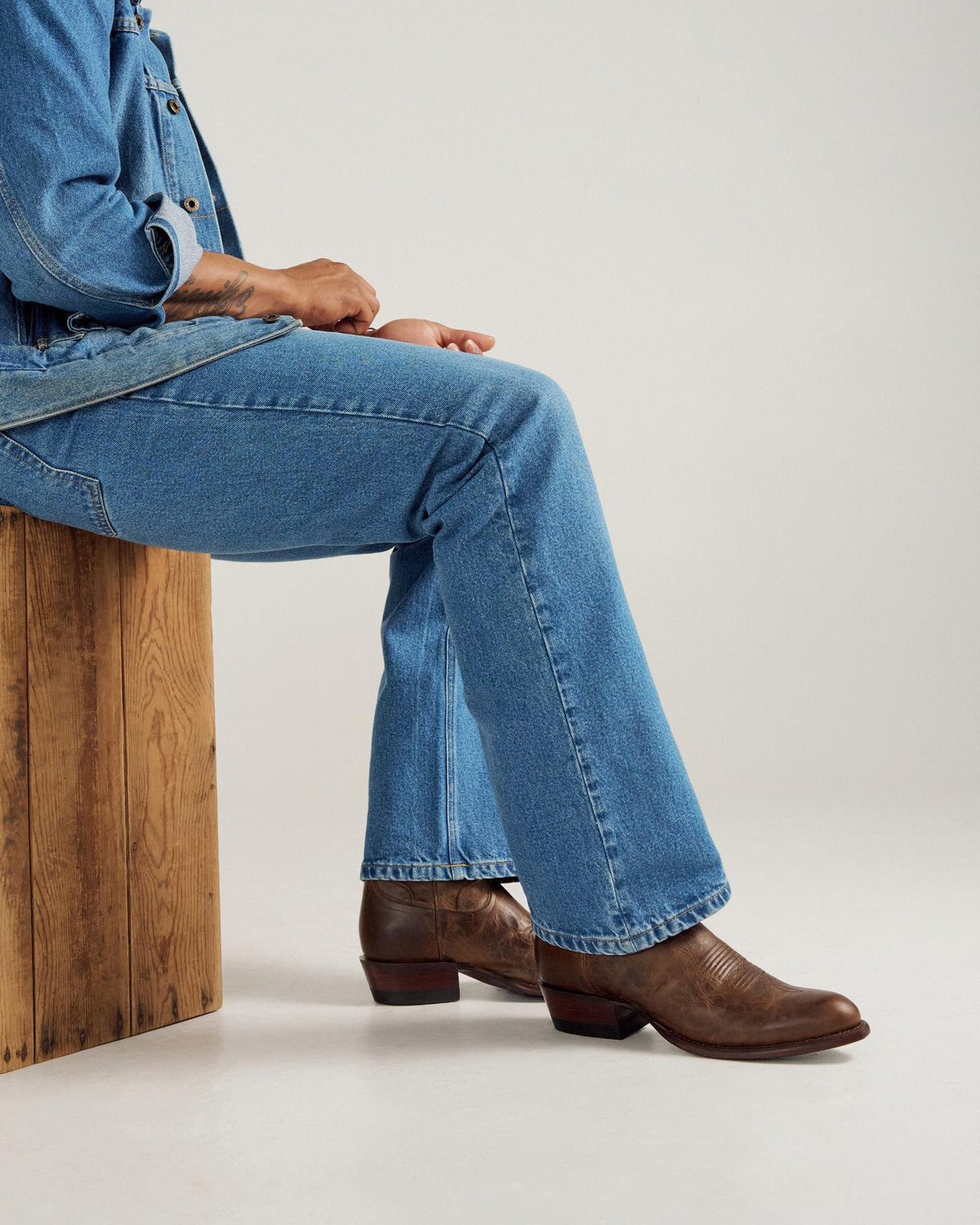 A person wearing blue jeans and brown leather boots sits on a wooden box against a plain background.
