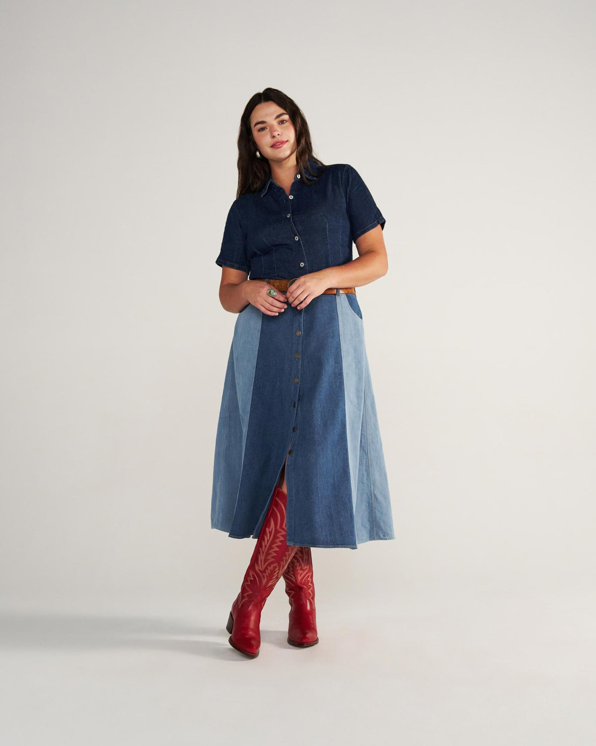 The Abby Wide Calf in Crimson Cowhide. Woman stands against a plain background wearing a short-sleeve denim shirt, two-tone button-front denim skirt, and striking crimson boots.