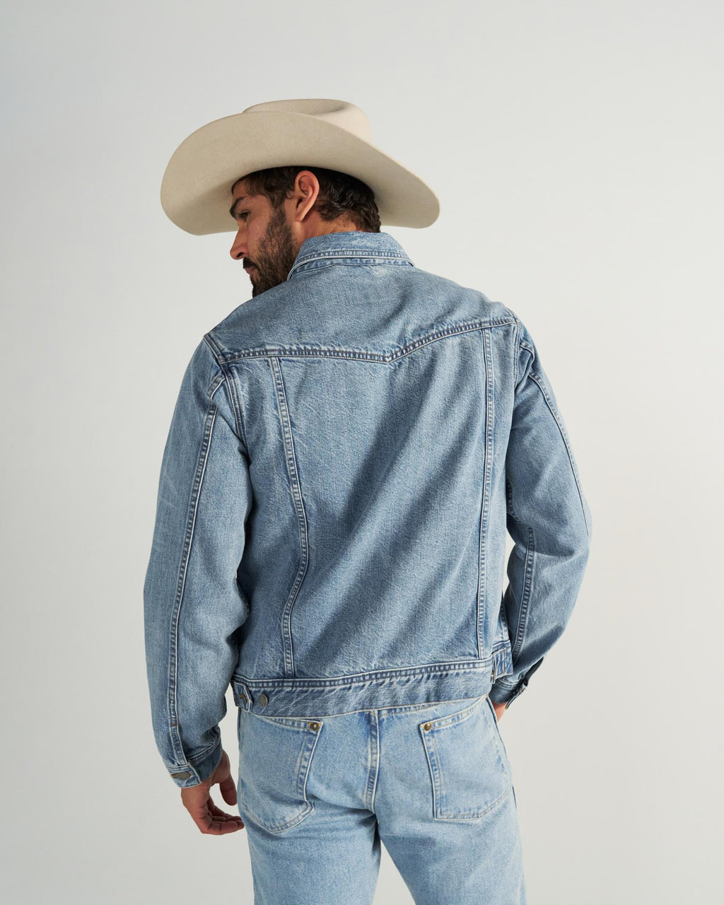 A man wearing a light denim jacket, matching jeans, and a beige cowboy hat stands facing away from the camera against a plain background.