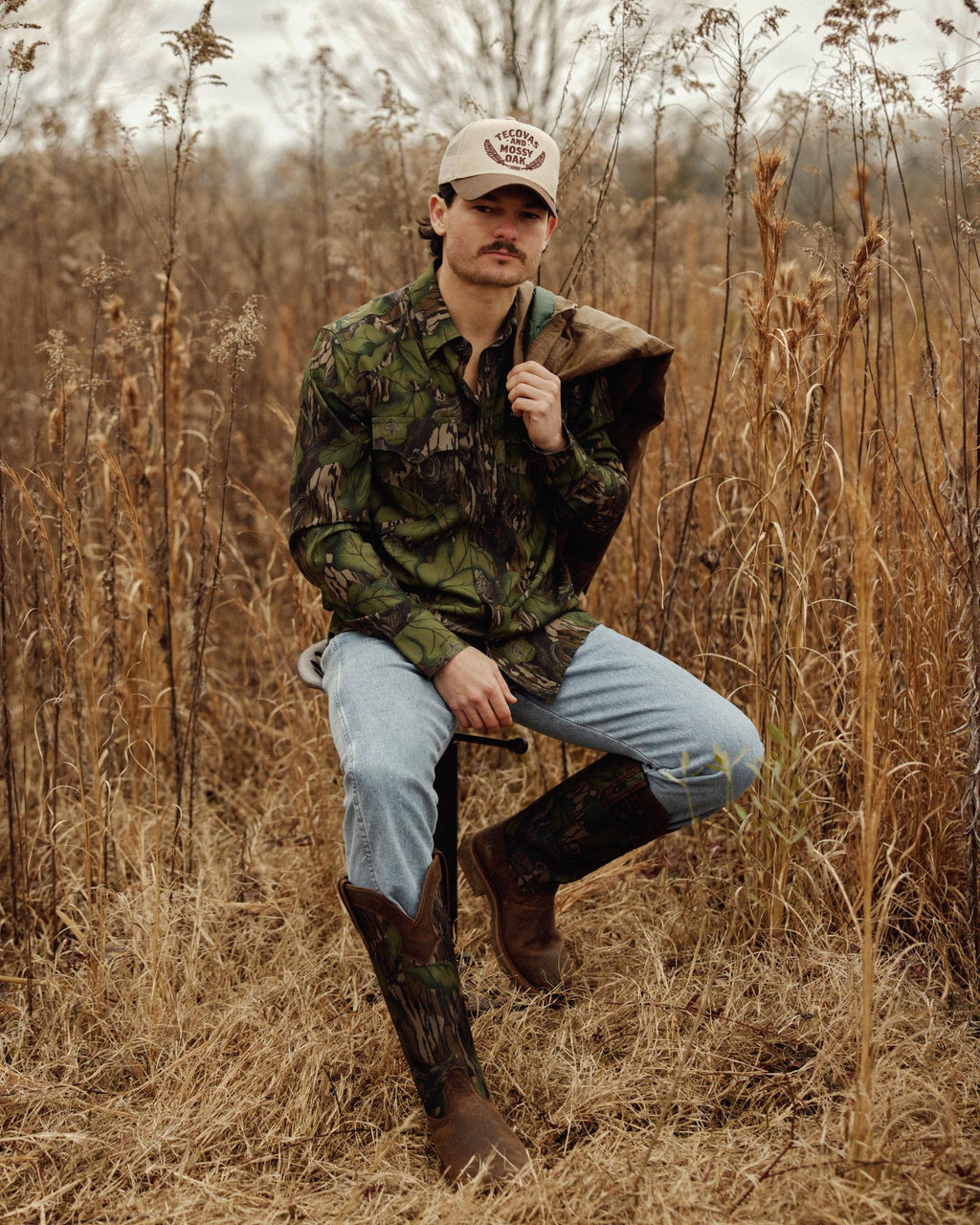 A man wearing a camouflage shirt, blue jeans, cowboy boots, and a beige cap sits on a stool in a dry grassy field, holding a jacket over his shoulder.