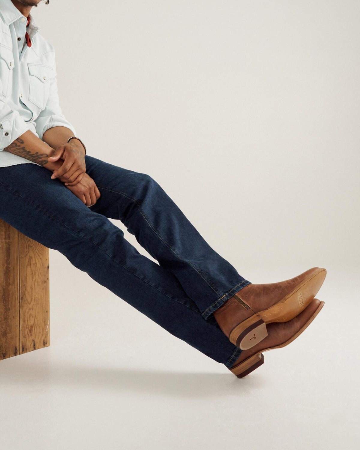 A person in a light shirt and blue jeans sits on a wooden bench, legs crossed, wearing brown cowboy boots.