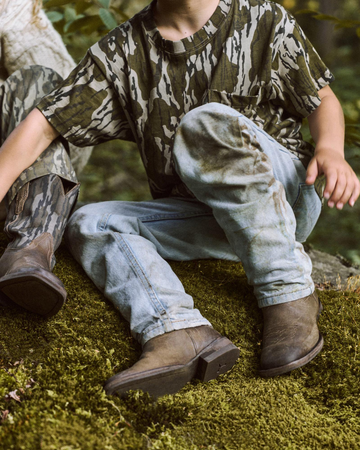 Two children sit on mossy ground outdoors, wearing camouflage shirts, light blue jeans, and brown boots. Only their bodies and partial faces are visible.