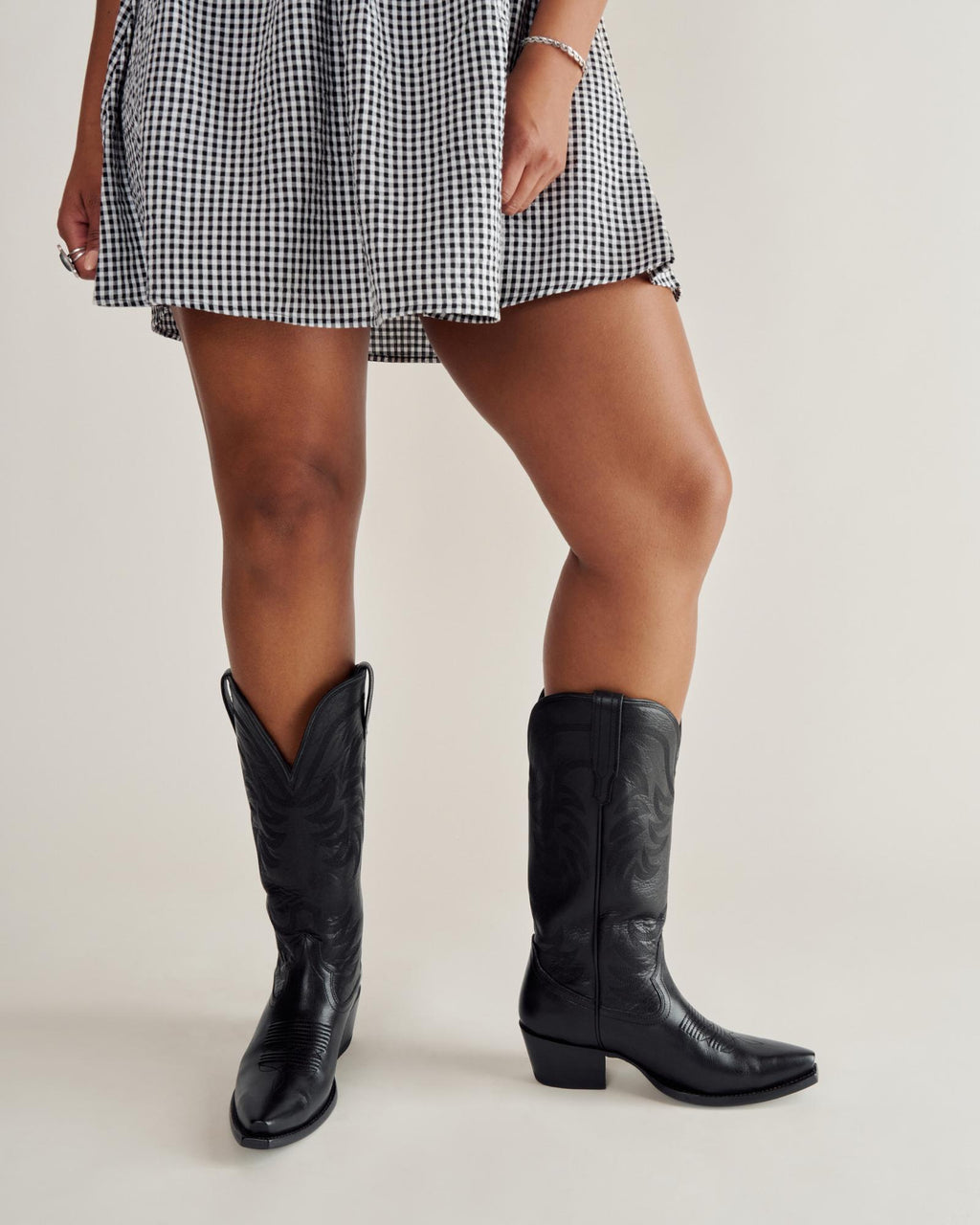A person wearing a black and white checkered skirt and black cowboy boots stands against a plain background.