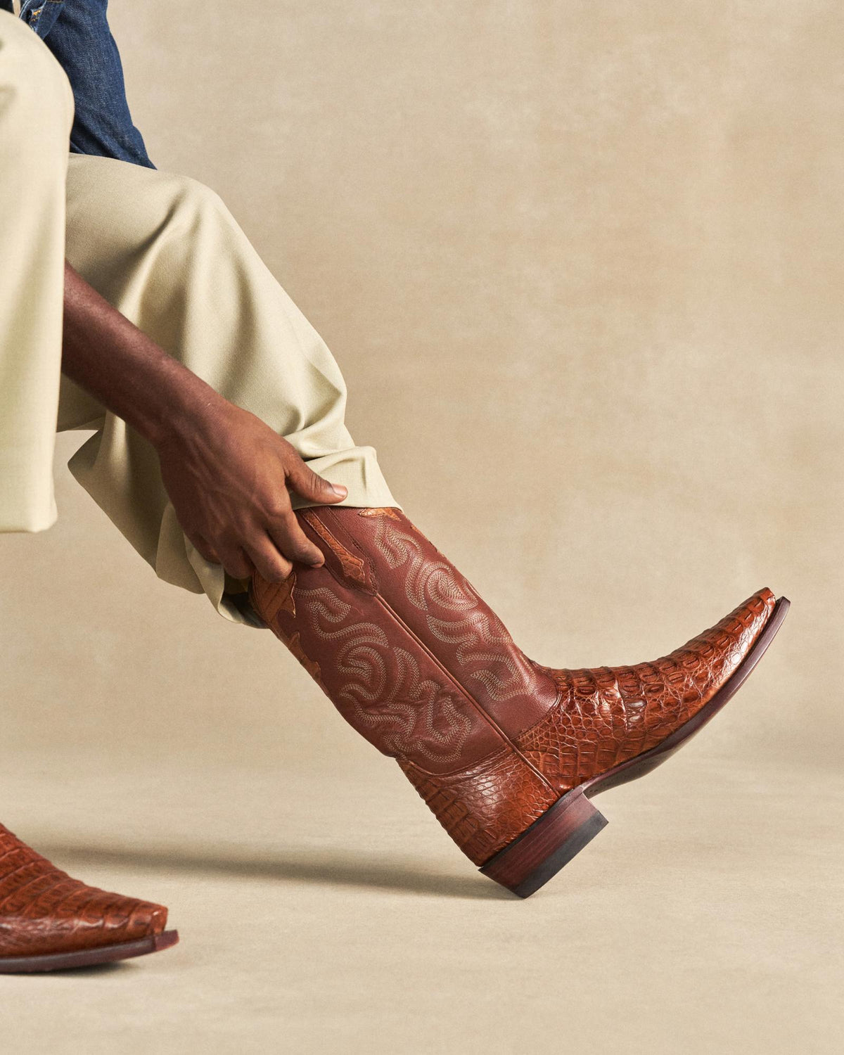 The Garrett in Pecan Hornback Caiman. Person wearing light pants pulls on a brown, patterned Pecan Hornback Caiman cowboy boot with a pointed toe.