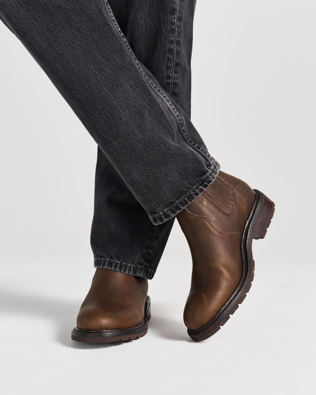 The Chelsea in Cafe Cowhide. Person wearing dark denim jeans and brown leather boots from The Chelsea Cowhide stands on a white background with one leg crossed in front of the other.