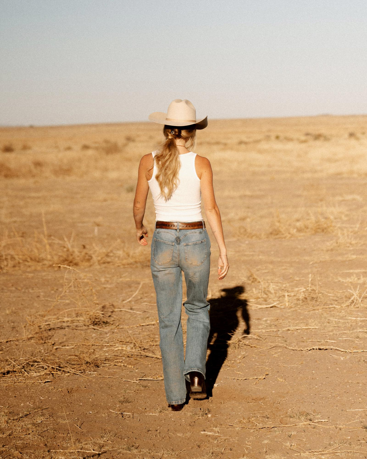 A person wearing a cowboy hat, white tank top, and blue jeans walks across a dry, open field with sparse vegetation.