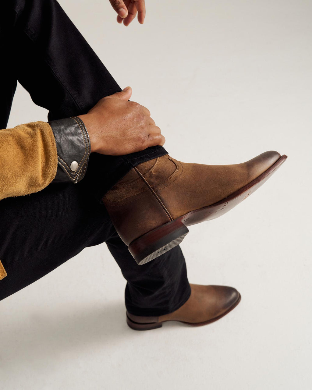A person wearing black pants and brown leather boots sits with one leg crossed over the other, showing the side of the boot and a tan jacket sleeve.