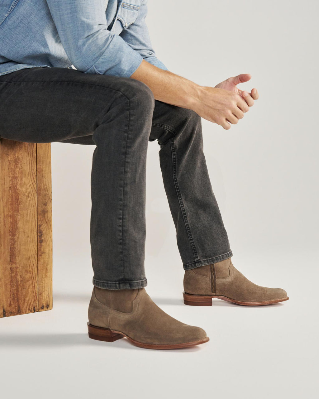 The Dean in Granite Suede. A man sitting on a wooden bench wears a light blue shirt, black jeans, and The Dean Suede Chelsea boots in taupe.