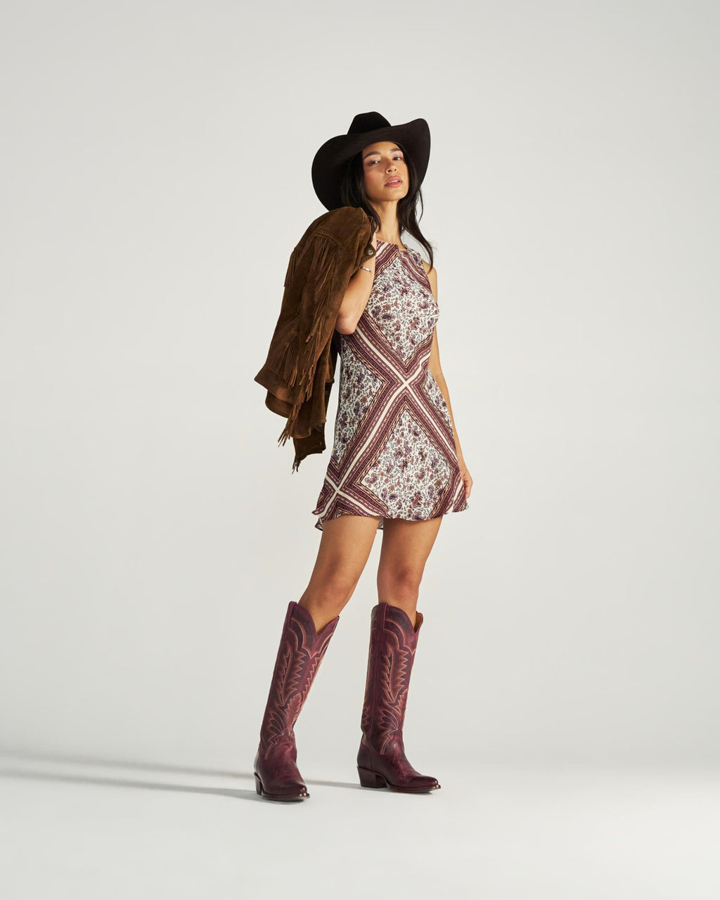 The Abby in Wine Goat. A woman in a cowboy hat, patterned dress, and boots stands holding a brown fringed jacket over her shoulder against a plain background, capturing the free-spirited style of The Abby Goat.