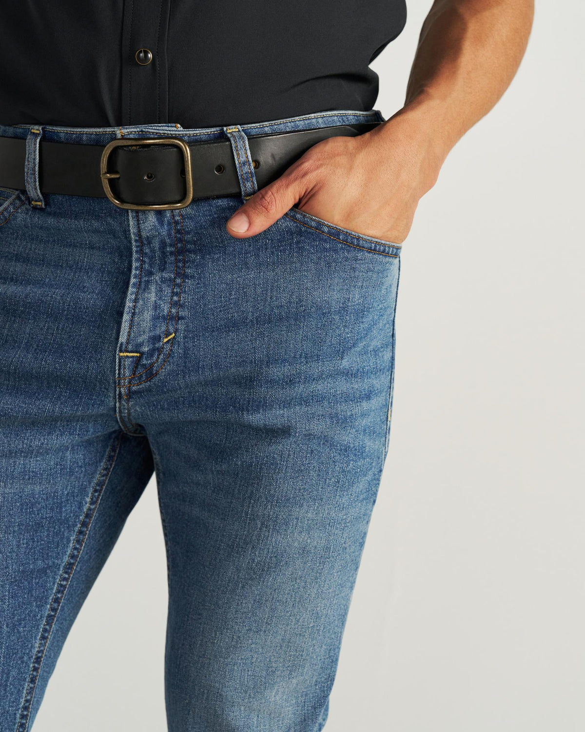 Person wearing blue jeans, a black shirt, and a black belt with a gold buckle, with their right hand in their pocket.