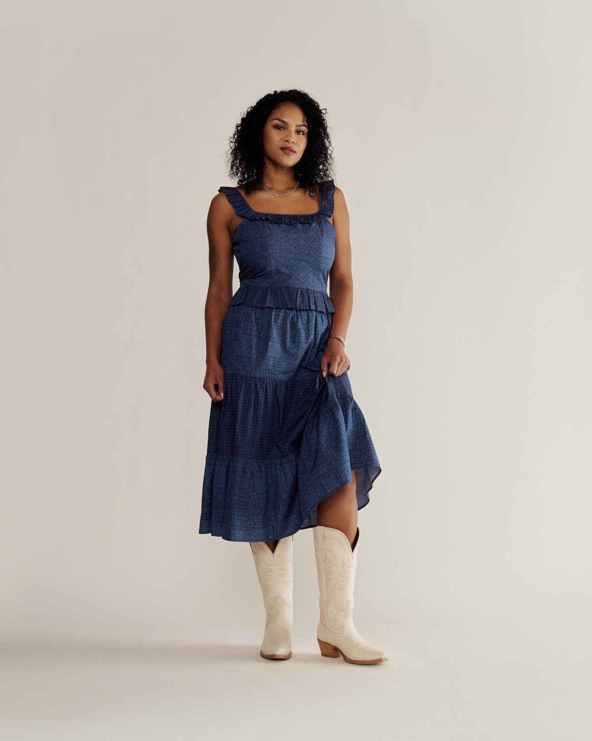 A woman in a blue sleeveless dress and white cowboy boots stands against a plain beige background, looking at the camera.