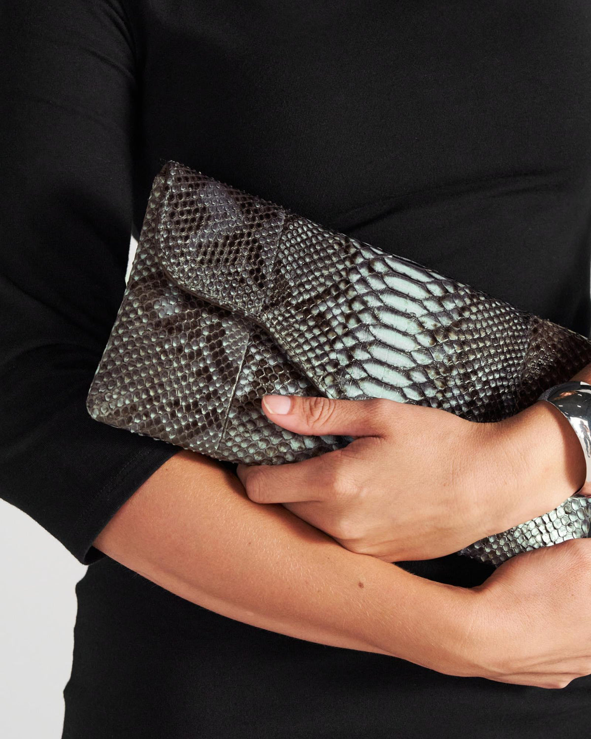 A person in a black long-sleeved top holding a grey and black snakeskin-patterned clutch.