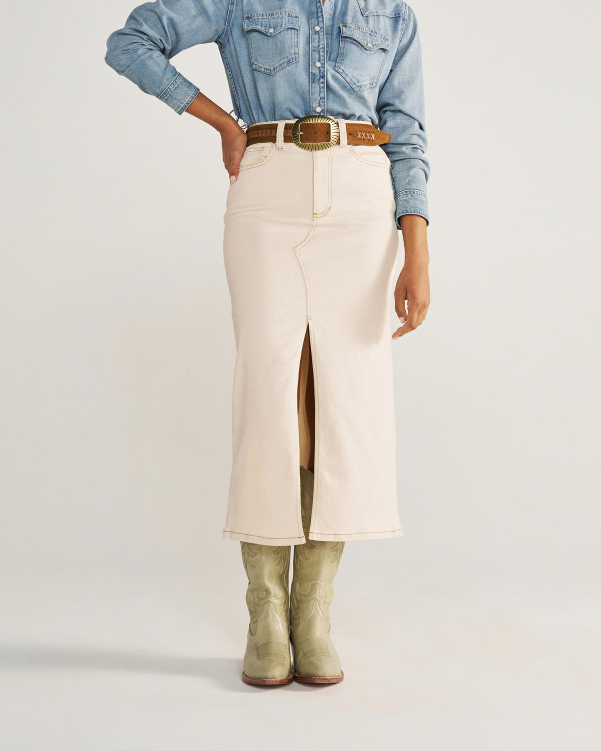 Person wearing a light denim shirt, cream-colored skirt with a front slit, tan belt with a large buckle, and light green cowboy boots.