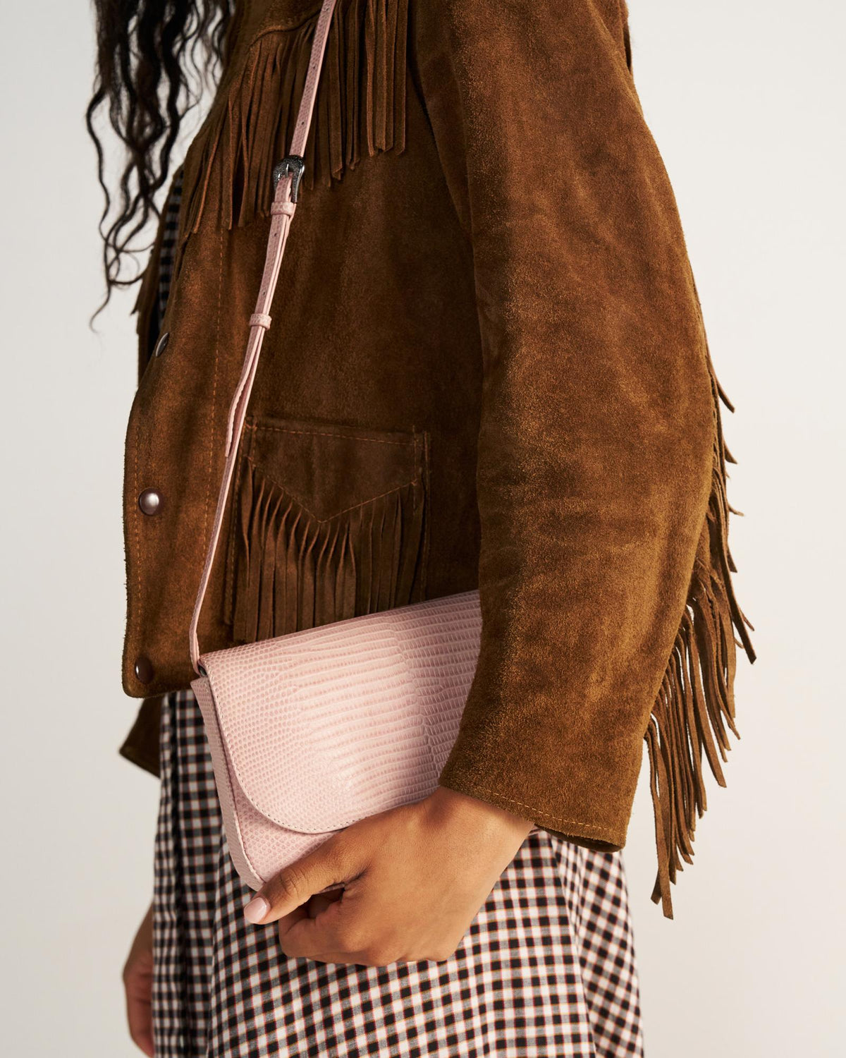 A person wearing a brown suede fringe jacket and a checkered dress holds a light pink crossbody bag.