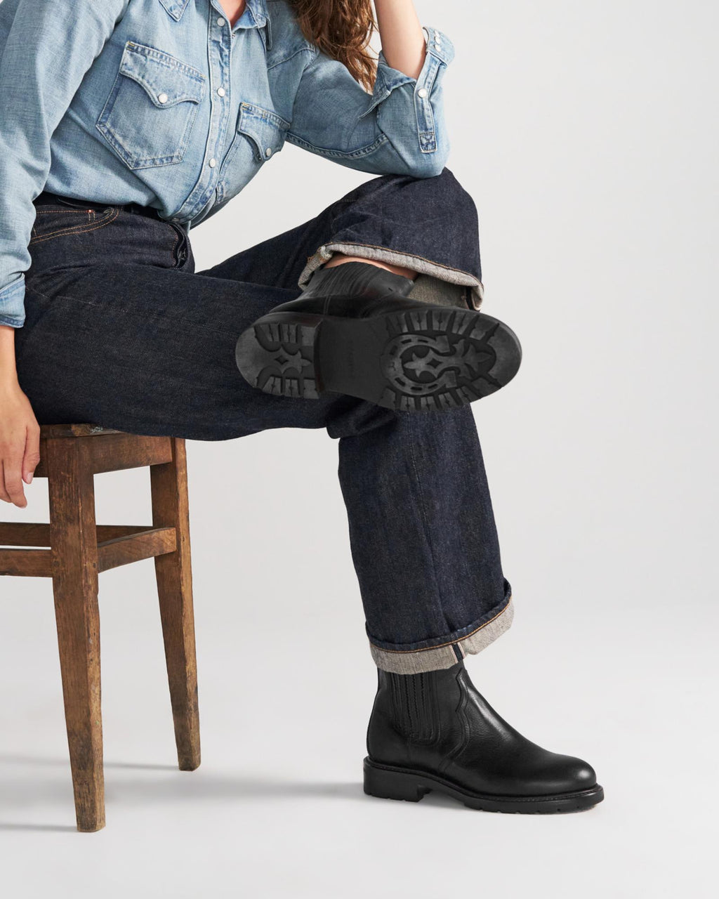 The Chelsea in Midnight Cowhide. A person in a denim shirt and jeans sits on a wooden chair with one leg crossed, showing the sole of their black Midnight Cowhide boot.