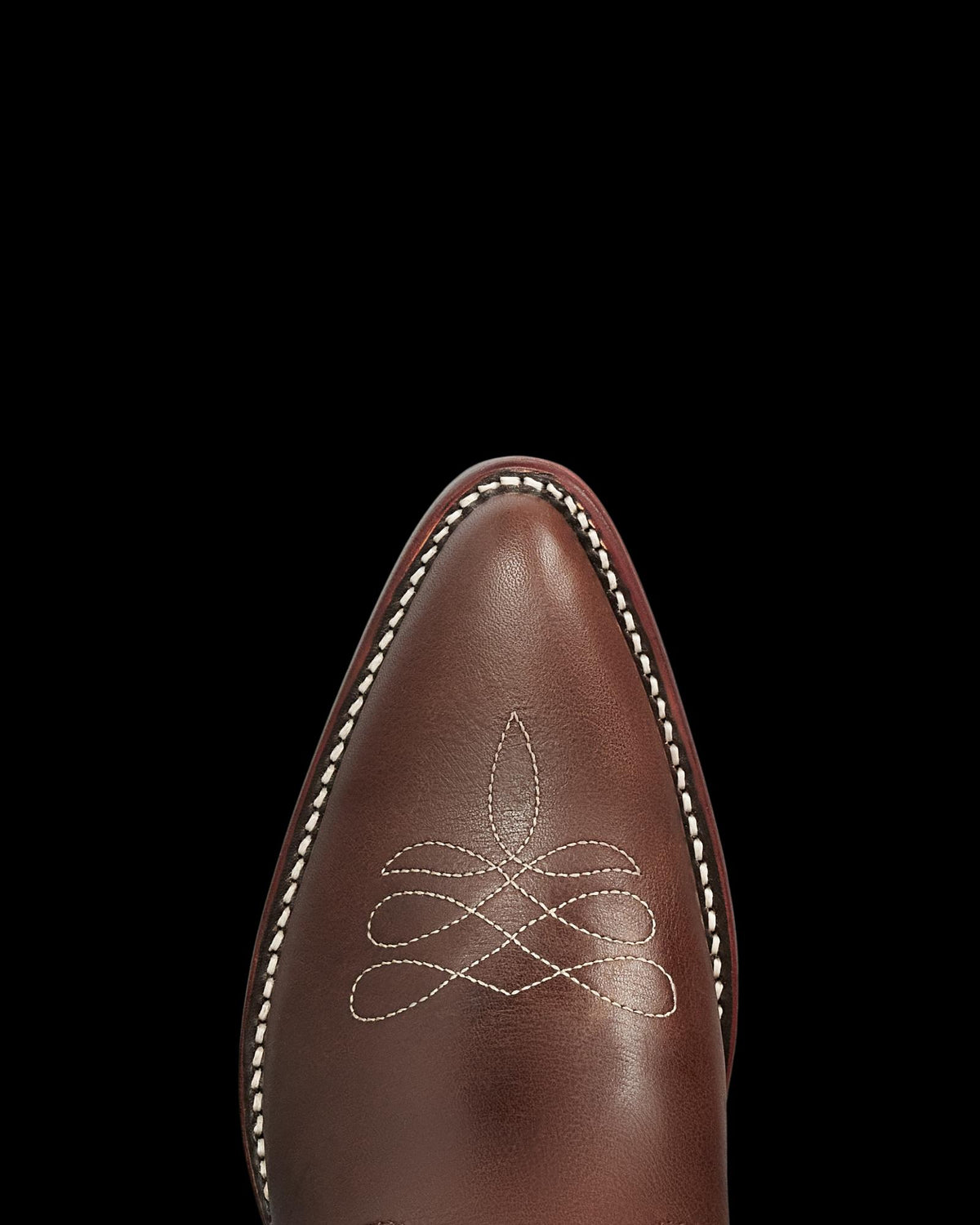 The Jolene in Truffle Cowhide. Close-up of the pointed toe of The Jolene Cowhide brown leather cowboy boot with white decorative stitching and a white-stitched welt, against a plain background.